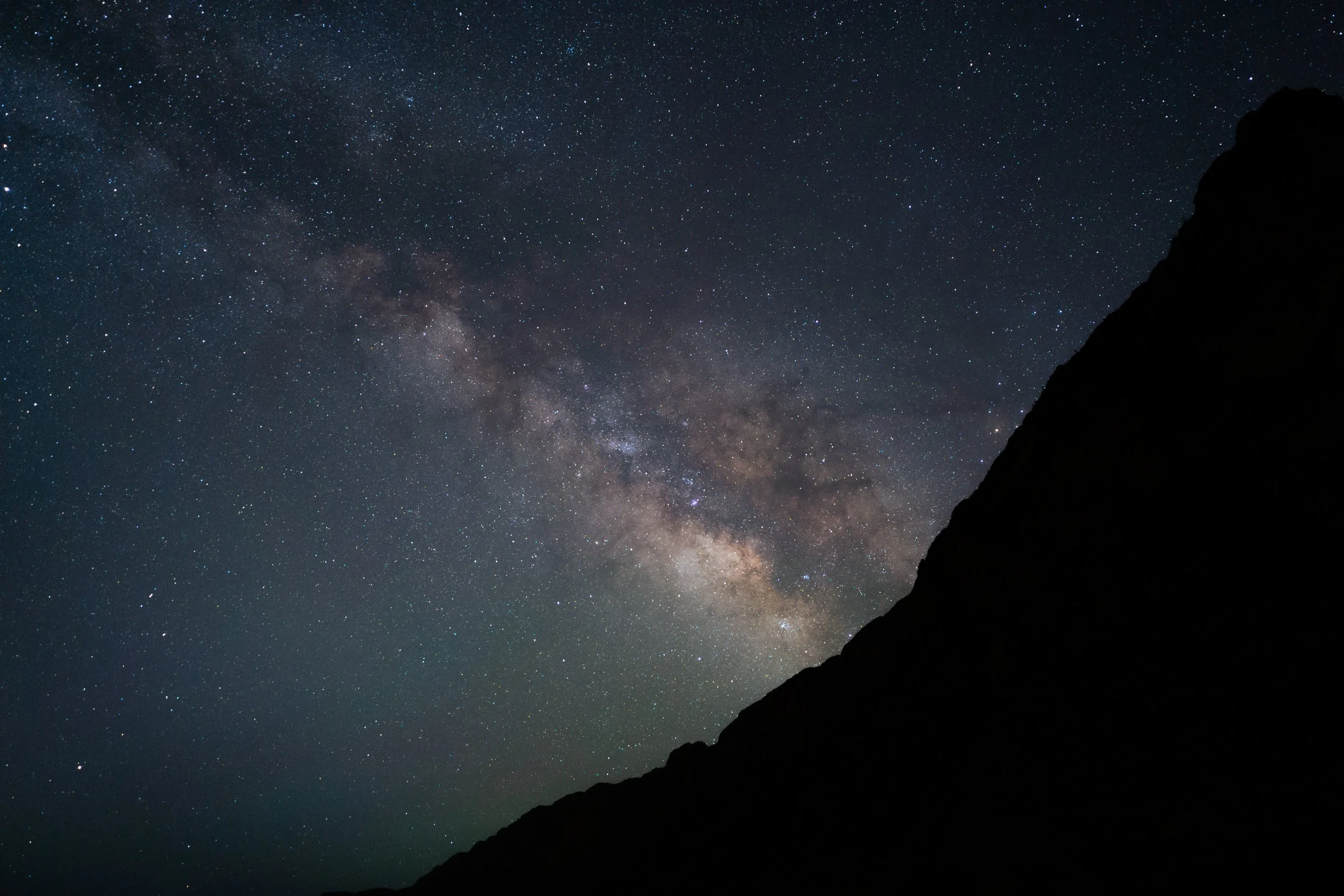 Nighttime sky filled with stars and faint Milky Way galaxy, silhouetted mountain in the foreground.