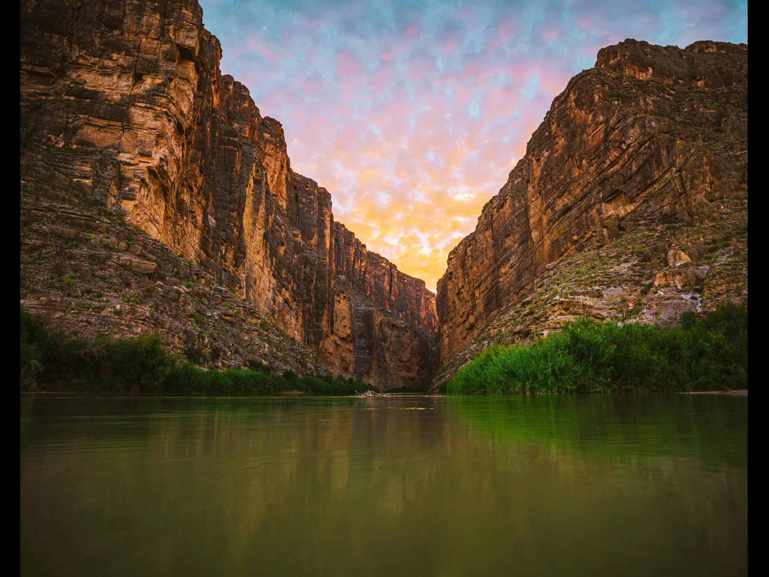Scenic view of tall, rugged rock canyon walls on either side with a grassy area at the base, and a calm river flowing through the bottom, with a colorful sky during sunset or sunrise in the background.