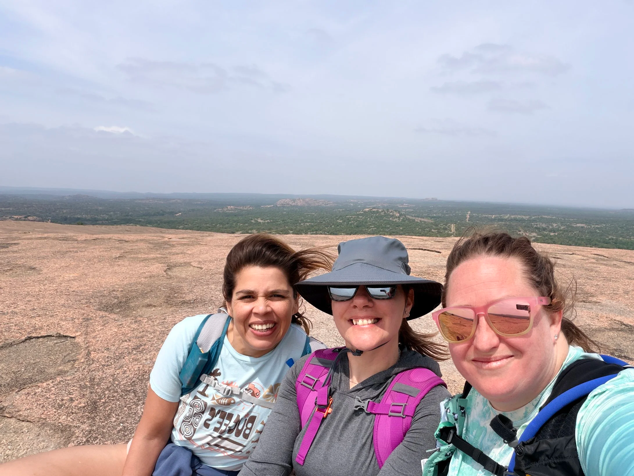 Group of four diverse women taking a selfie outdoors on a rocky hilltop with a vast landscape and cloudy sky in the background.
