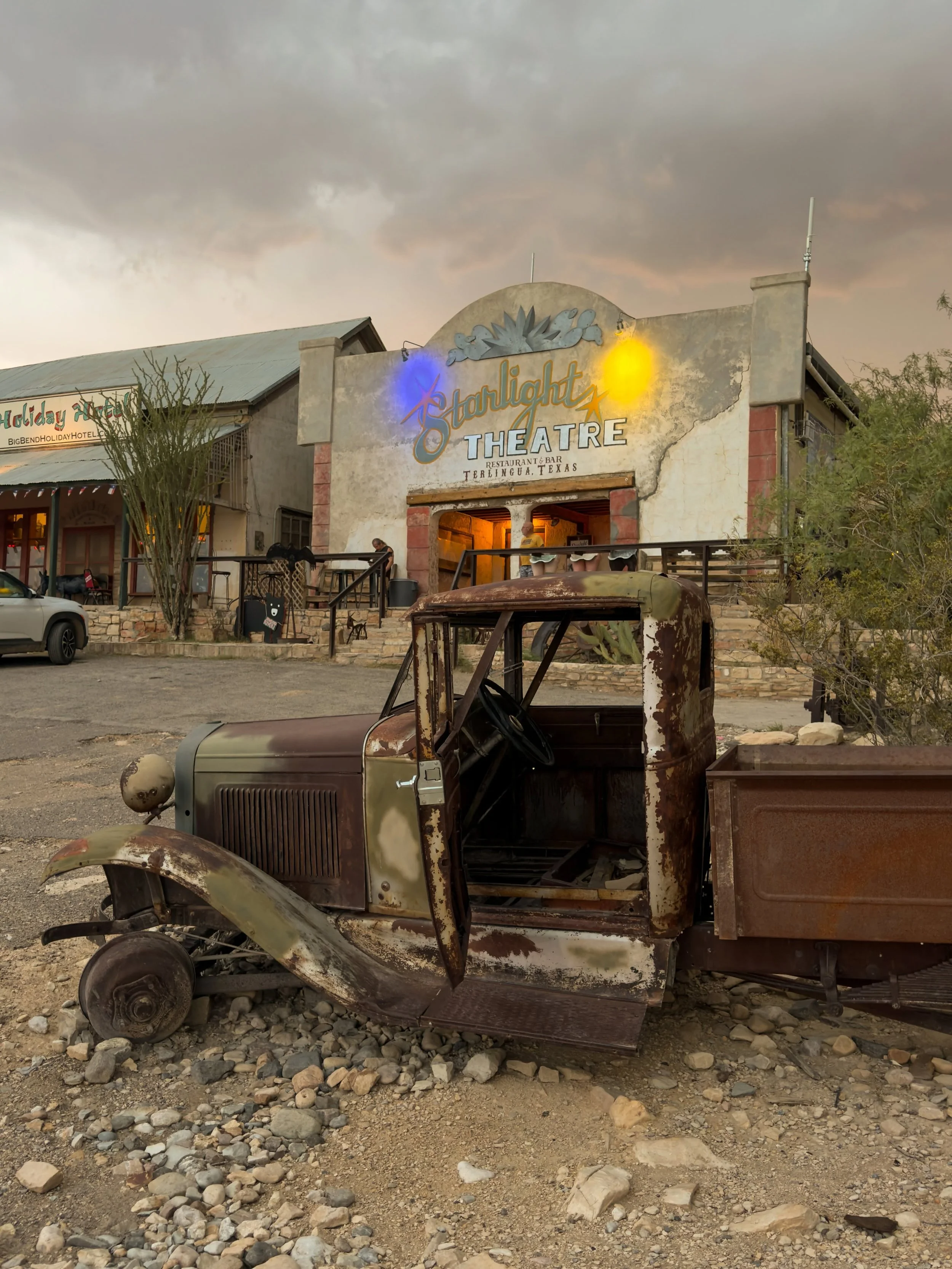 A rusty, vintage pickup truck with missing wheels parked in front of a rustic building with a sign reading 'Starlight Theatre' in Terlingua, Texas. The building has a weathered facade and neon lights, with a partly cloudy sky overhead.
