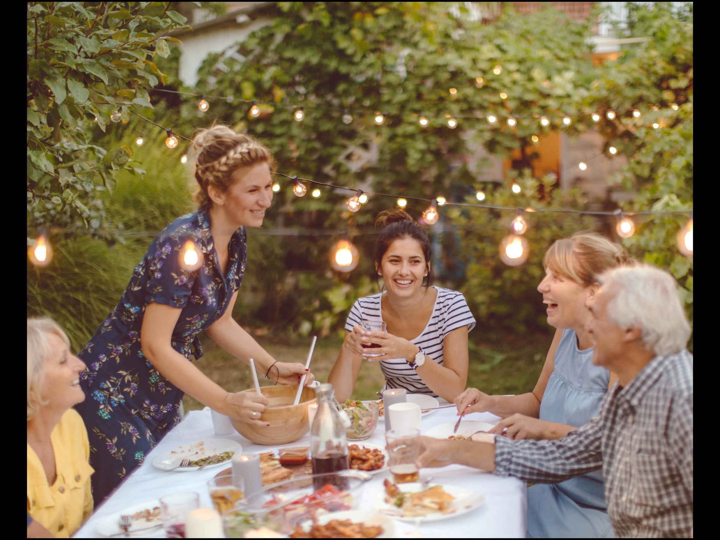 People enjoying an outdoor gathering around a table set with food, drinks, and a wooden bowl, under string lights in a backyard with trees.