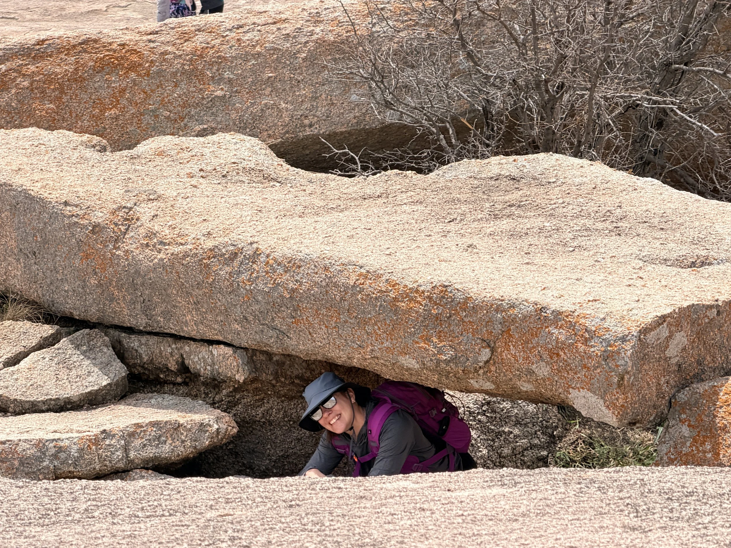 A woman smiling and wearing sunglasses, a hat, and a purple jacket, is crouching down under large reddish-brown rocks and small branches, with other hikers visible in the background.