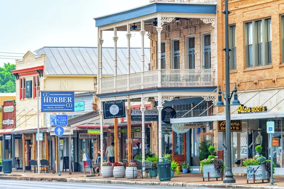 City street with historic buildings, retail stores, and outdoor seating, featuring a prominent two-story building with a balcony and decorative trim.