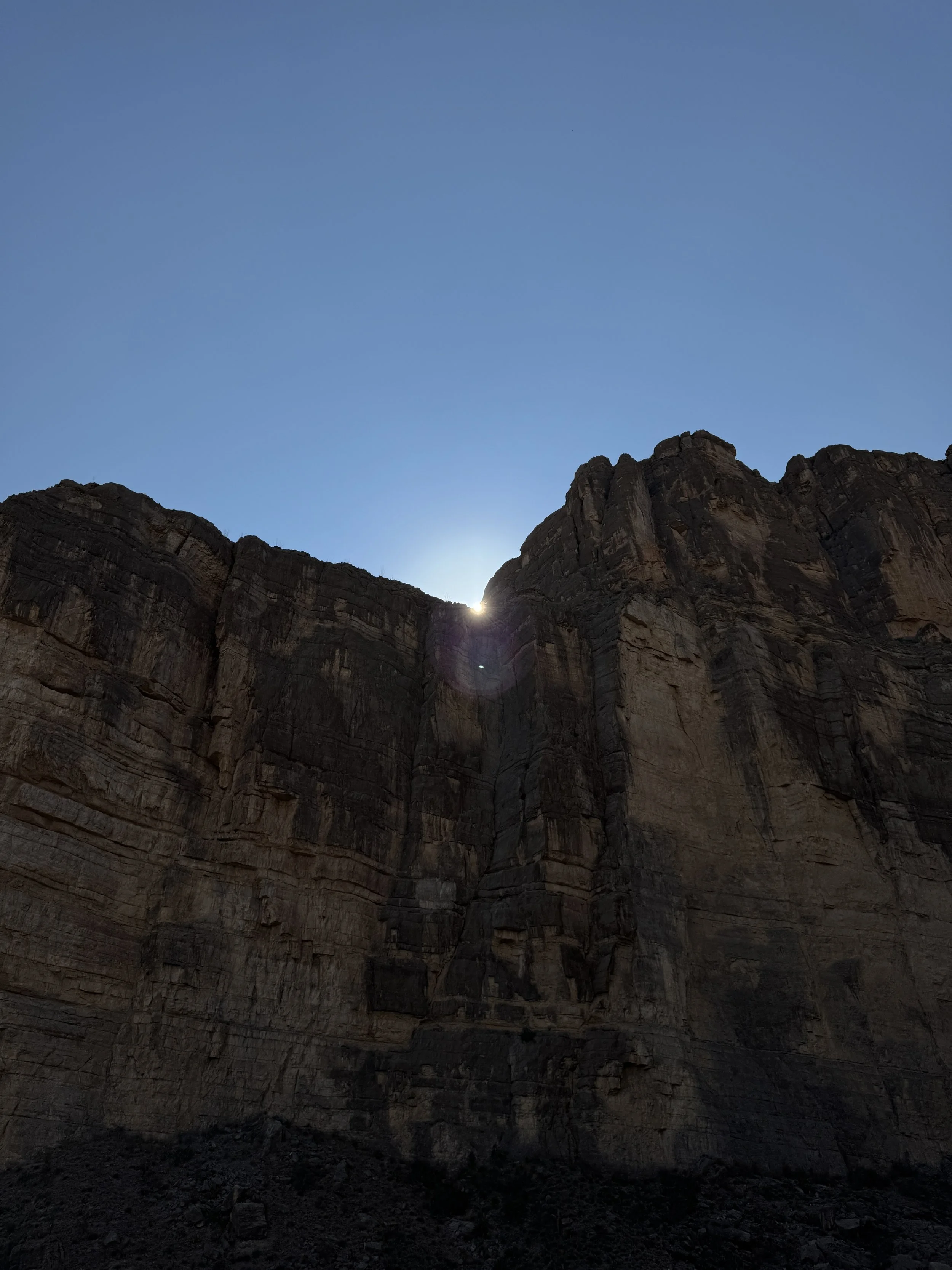 Sunset or sunrise behind tall mountain cliffs with clear blue sky.