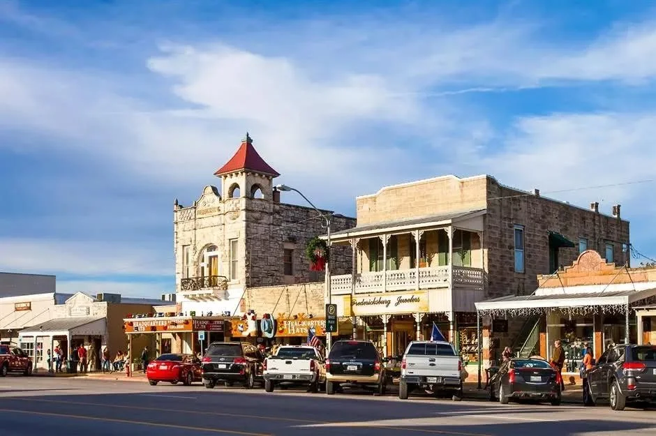 Historic buildings with shops and parked cars along a street under a blue sky.