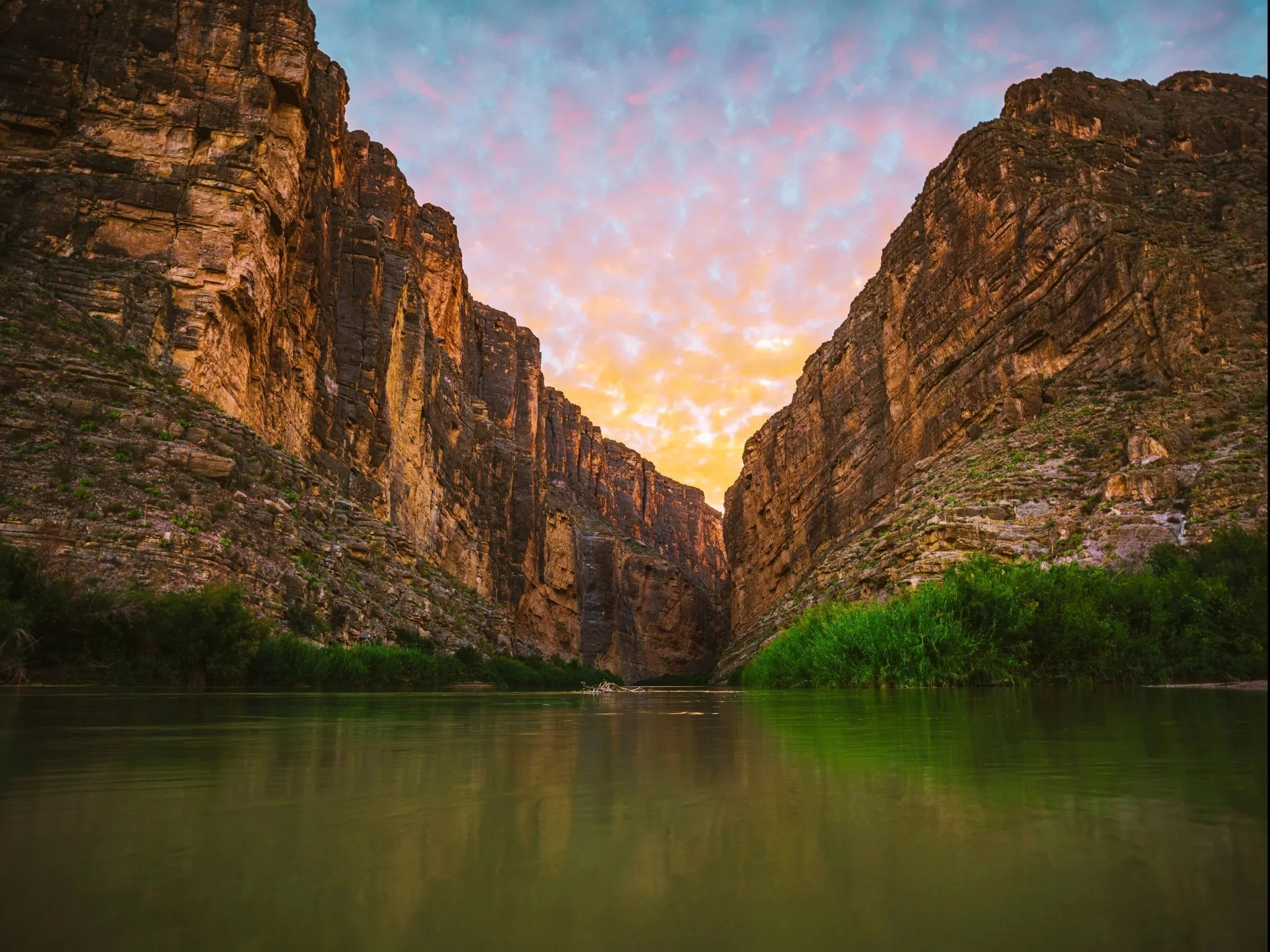 A canyon with steep rocky cliffs on each side, a calm river running at the bottom, and a colorful sky at sunset or sunrise with pink, orange, and blue hues.