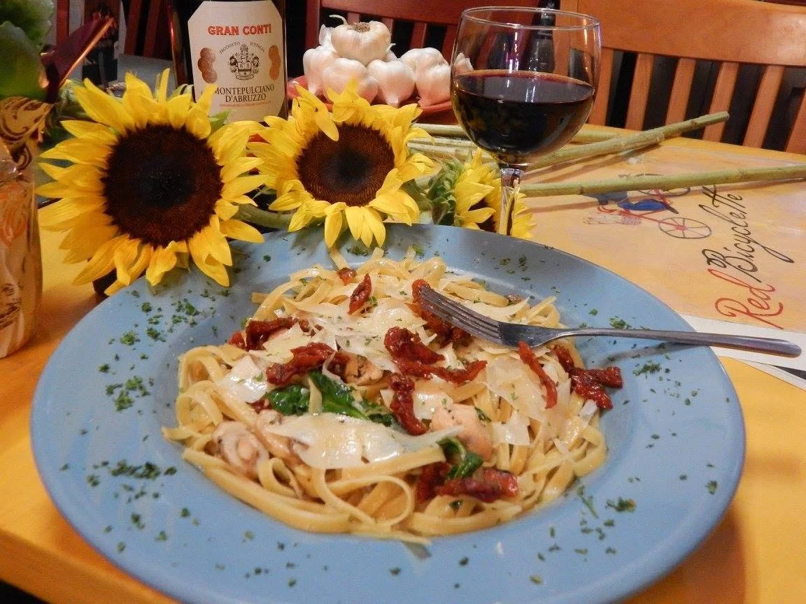 Plate of fettuccine pasta with sun-dried tomatoes, herbs, and cheese, on a table decorated with sunflowers, a glass of red wine, garlic, and a tablecloth with the words "Red Bicycle".