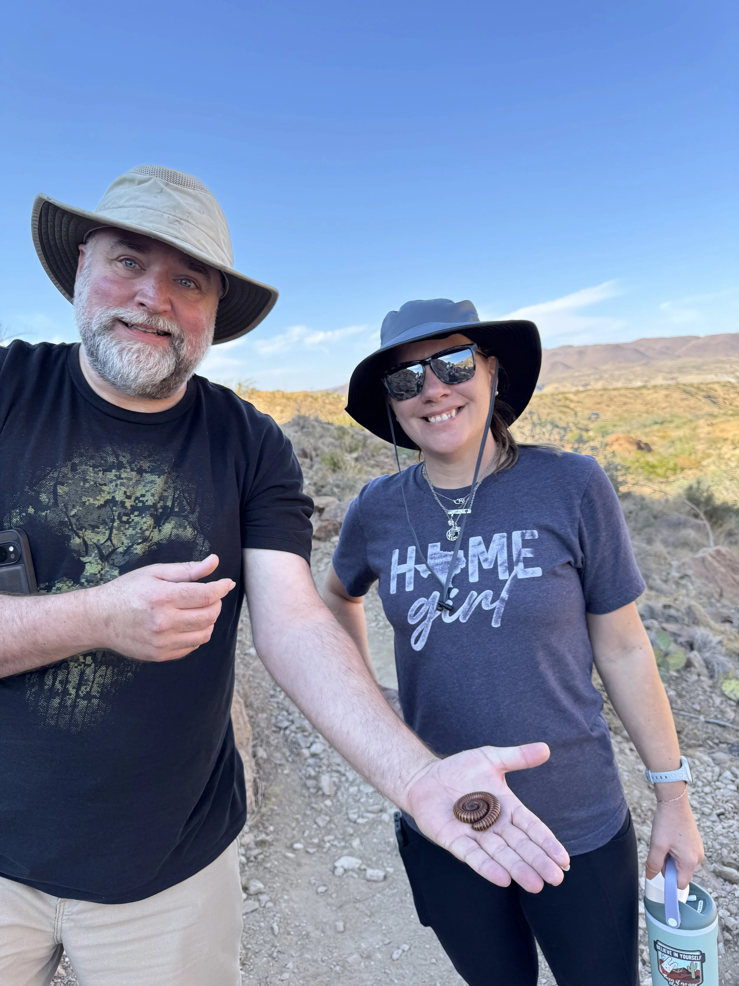 Two people outdoors, one showing a small desert millipede on their open hand, with desert landscape and mountains in the background.