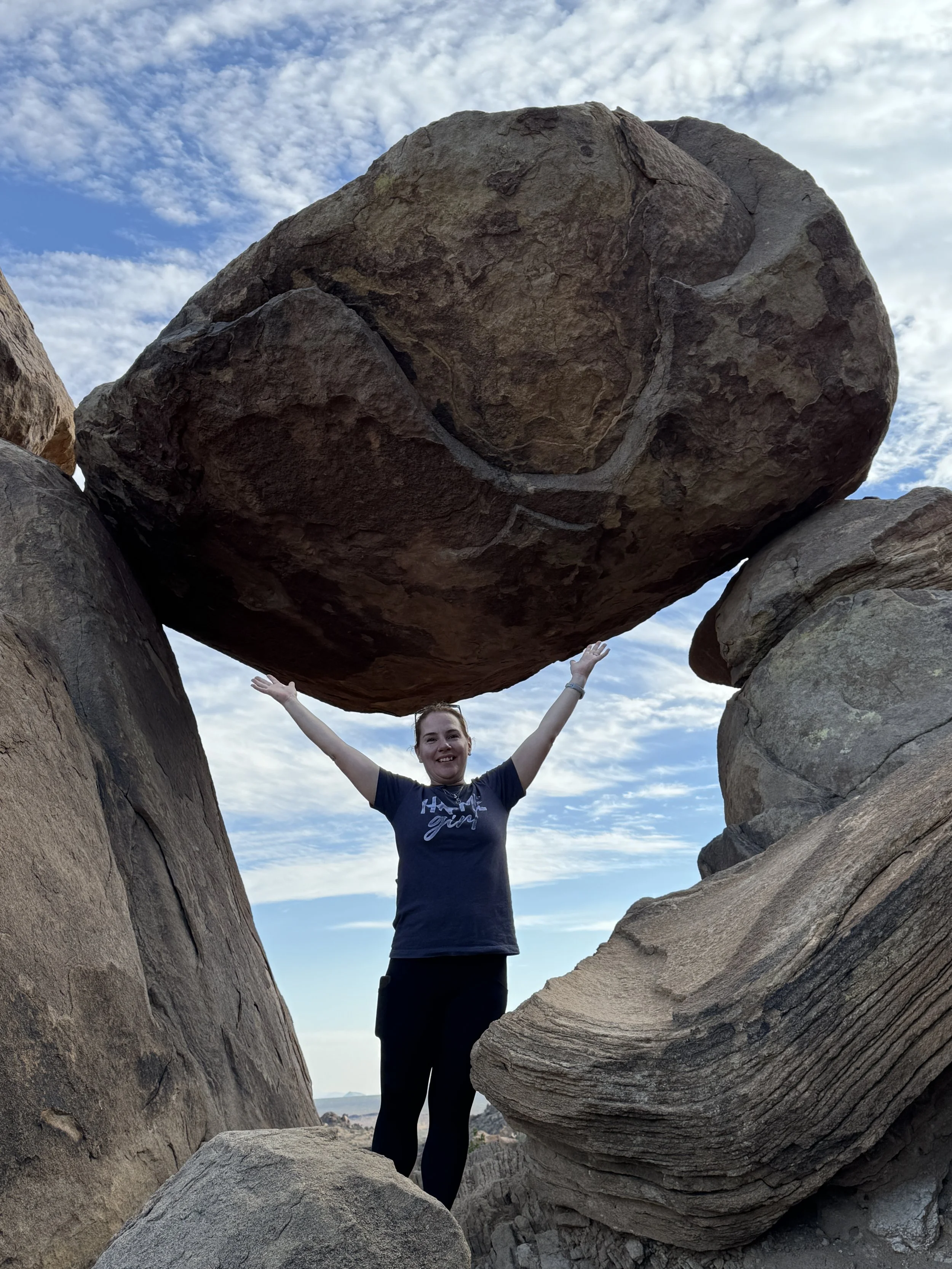 A woman standing between large rocks outdoors with her arms raised, with a large boulder above her, under a partly cloudy sky.