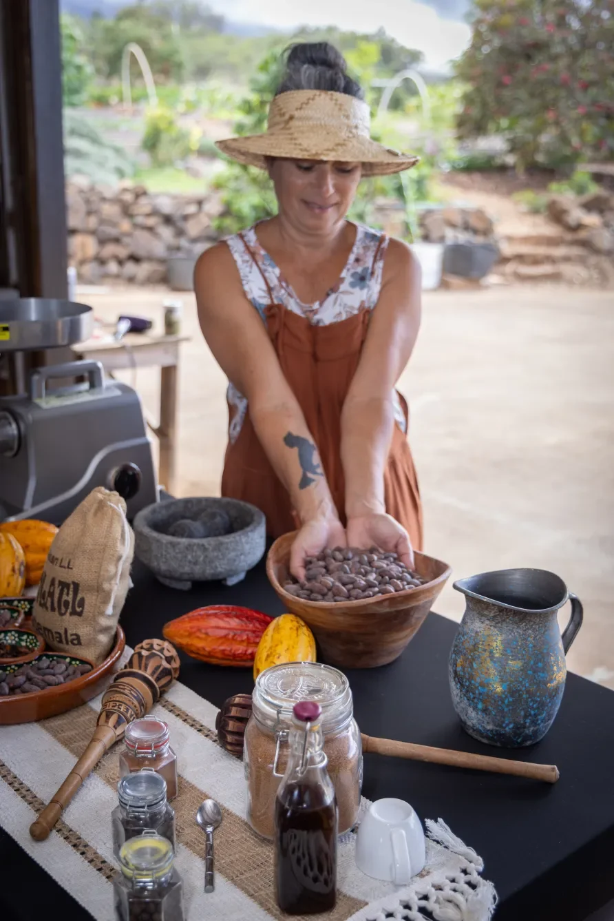 Woman wearing a straw hat and apron holding a bowl of cacao beans at a table with various items, set outdoors with a garden in the background.