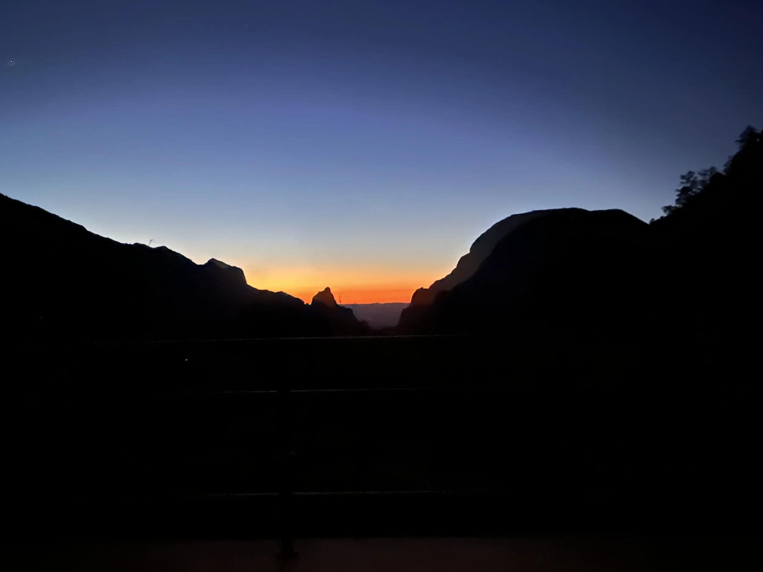A landscape photograph of a sunset or sunrise over a mountain valley with a clear sky, dark silhouette of mountains on either side, and warm light near the horizon.