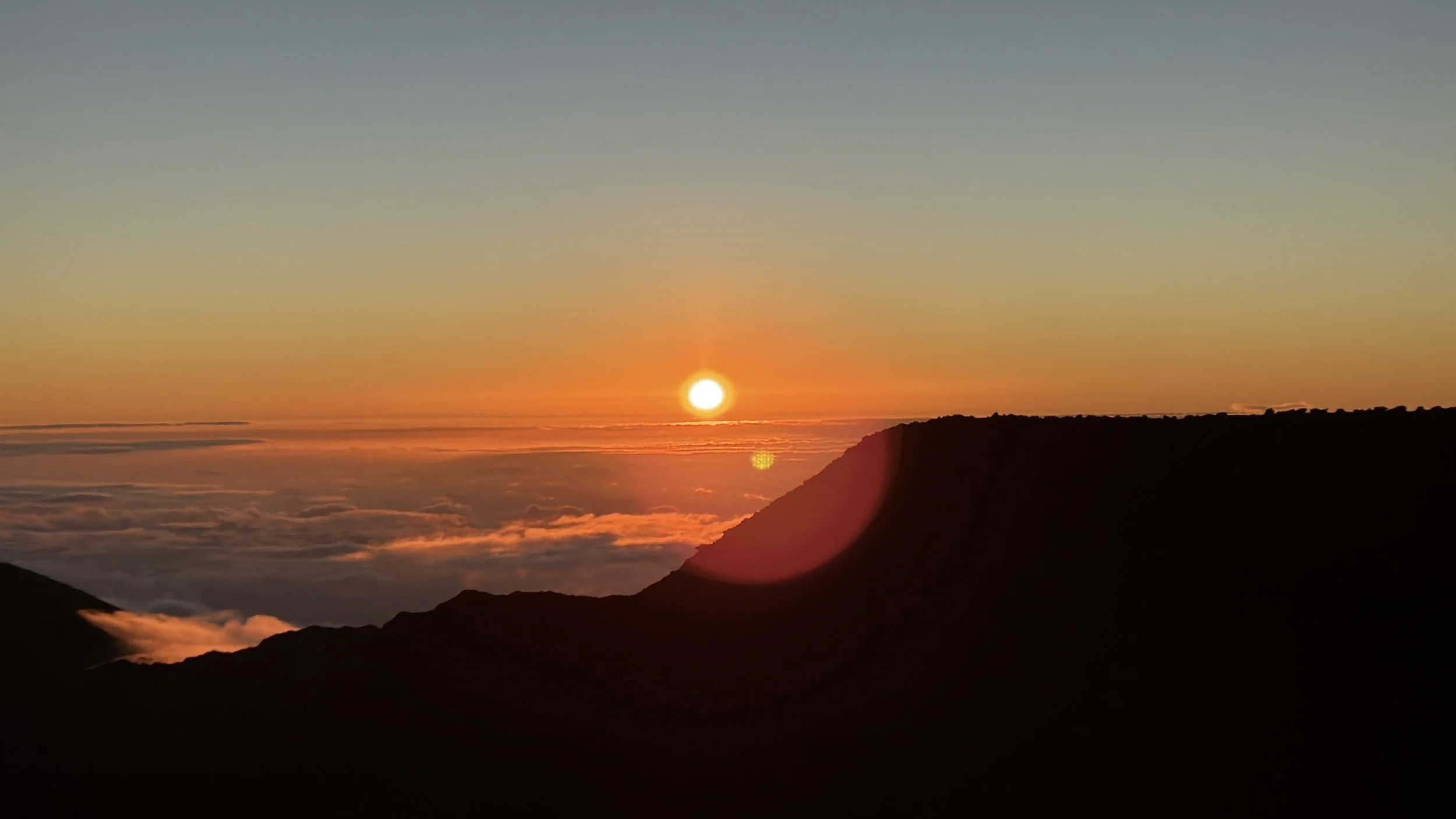 Sun setting over mountains with a colorful sky and clouds below.