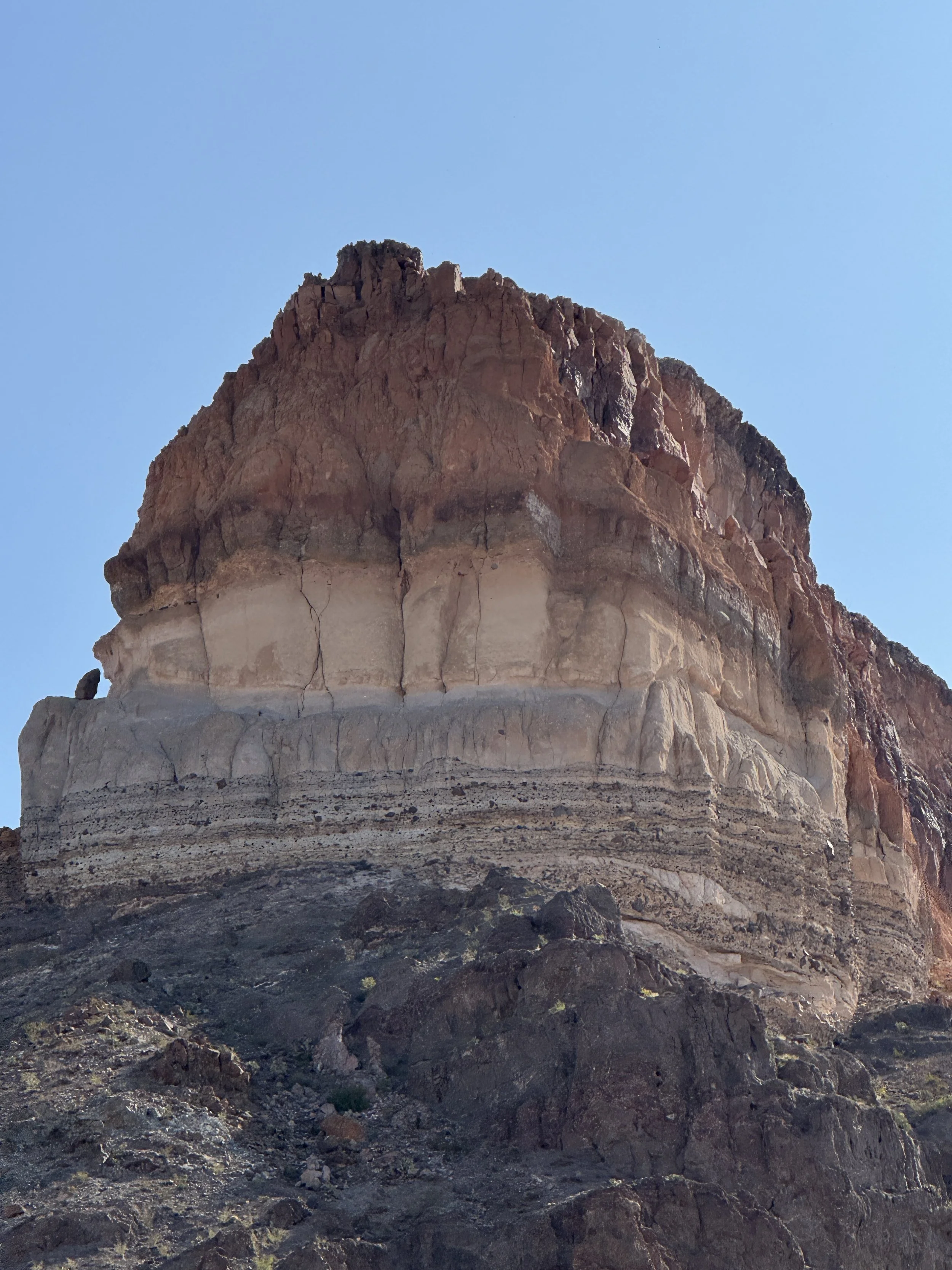 Close-up of a large, layered rock formation with a reddish-brown top against a clear blue sky.