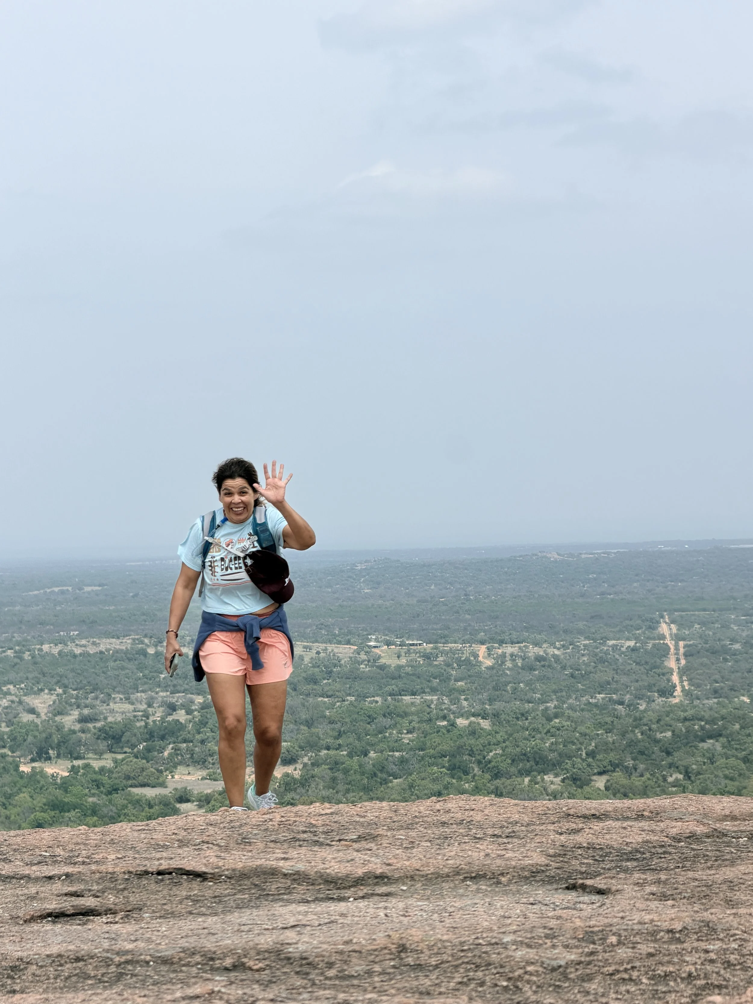 Woman standing on a rocky surface with a scenic landscape and cloudy sky in the background, waving and smiling.