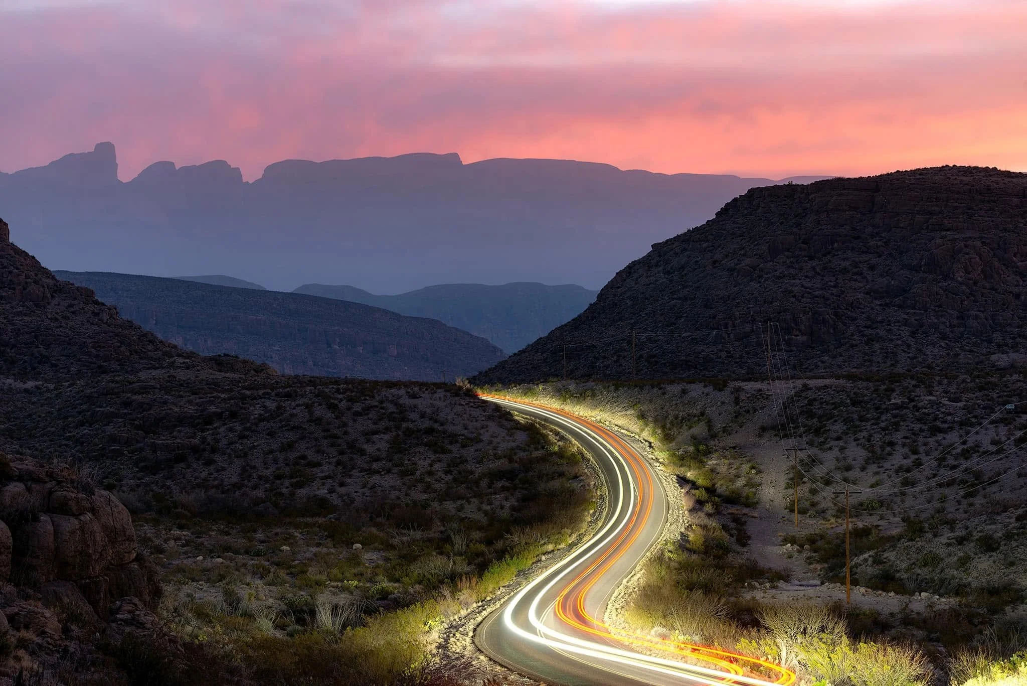 Long exposure photo of a winding desert highway at dusk with light trails from vehicles and mountain silhouettes in the background under a pink and purple sky.