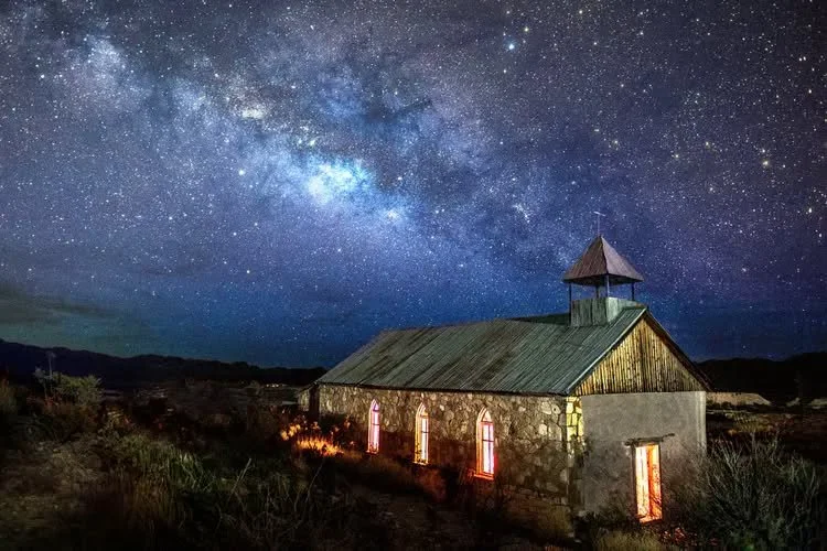 A stone church with a pitched roof and small tower under a starry night sky filled with the Milky Way galaxy.