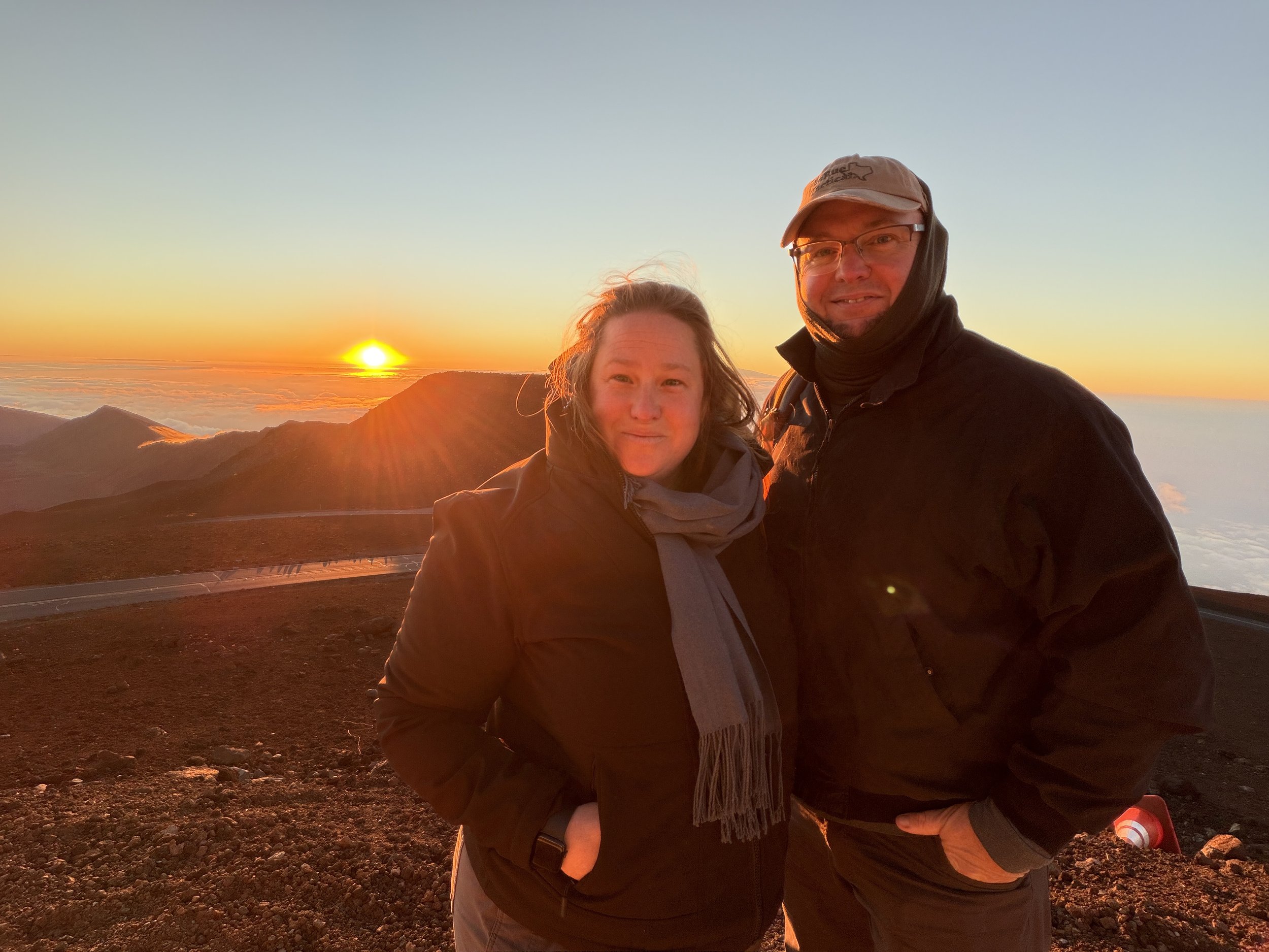 A man and a woman standing together outdoors during sunset or sunrise, with mountains and clouds in the background.