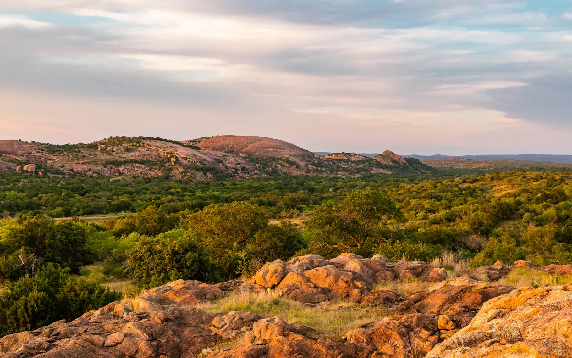 Landscape view of rolling hills and mountains with green forests and rocky terrain under a partly cloudy sky.