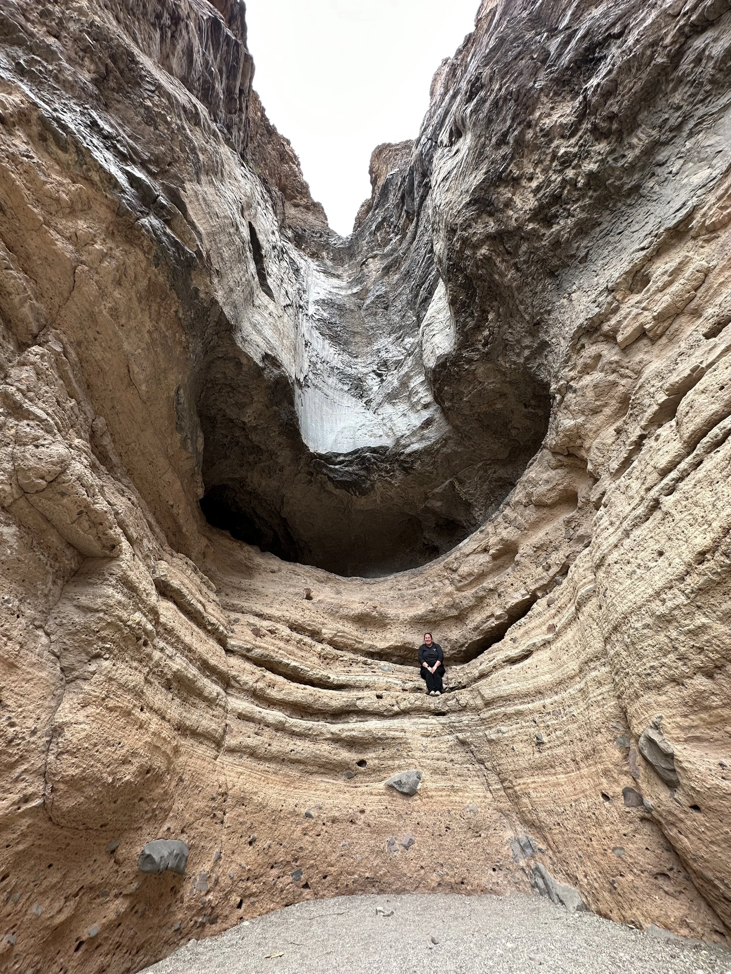 Person sitting inside a narrow canyon with layered, beige and brown rock walls.