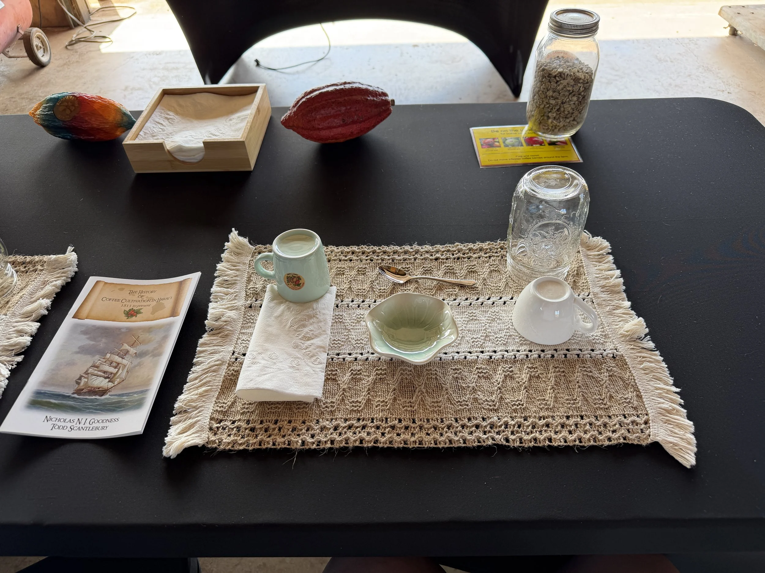 A table setting with a lace table runner, small white cups, a spoon, a green dish, a book titled "The History of Coffee Cultivation in Hawaii 1813 to Present," a paper bag, two decorative cocoa pods, a wooden box, and a jar of cereal with a yellow label.