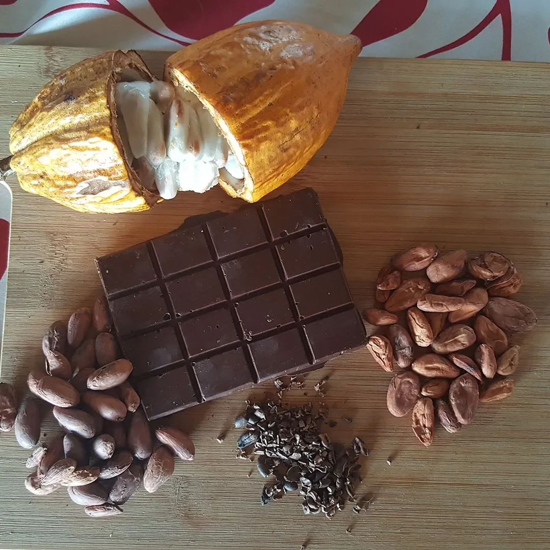 A halved roasted cocoa pod, a chocolate bar, and cocoa beans with their shells and chopped cacao nibs displayed on a wooden surface.