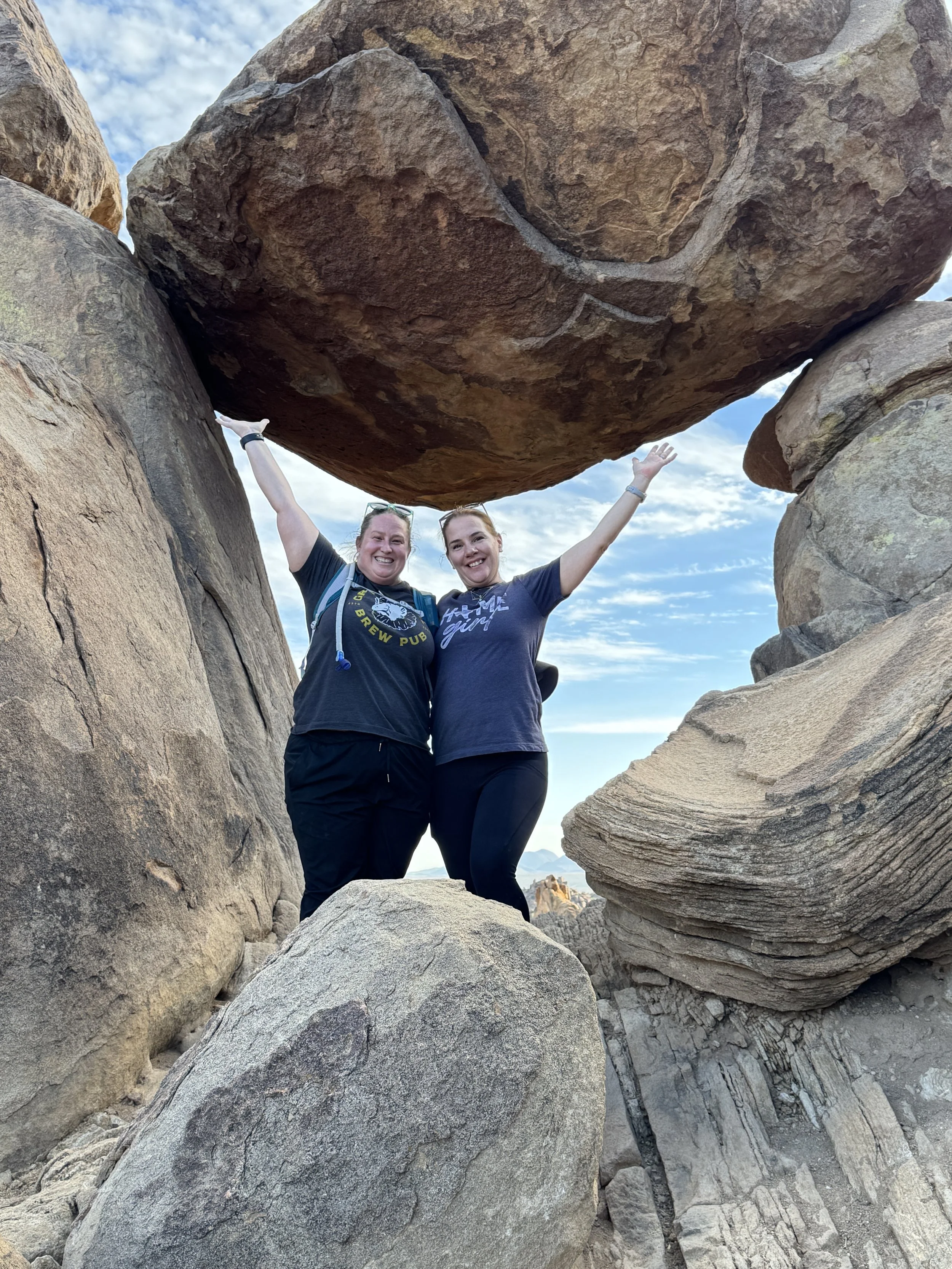 Two women smiling and raising their arms in front of large boulders and rock formations, during daytime with partly cloudy sky.