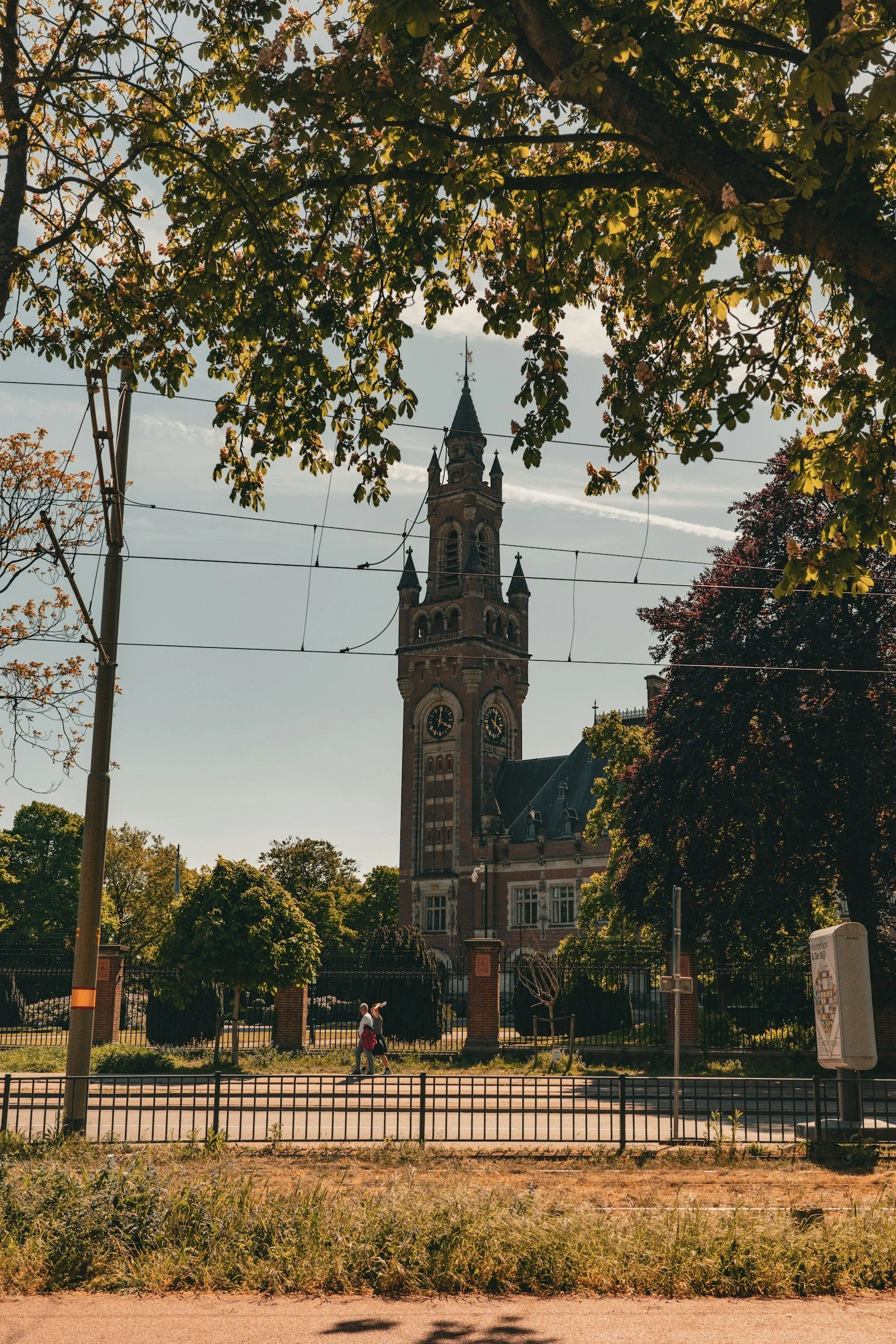 A historic brick clock tower with pointed turrets and a clock face, framed by leafy trees, with pedestrians walking on the sidewalk in front.