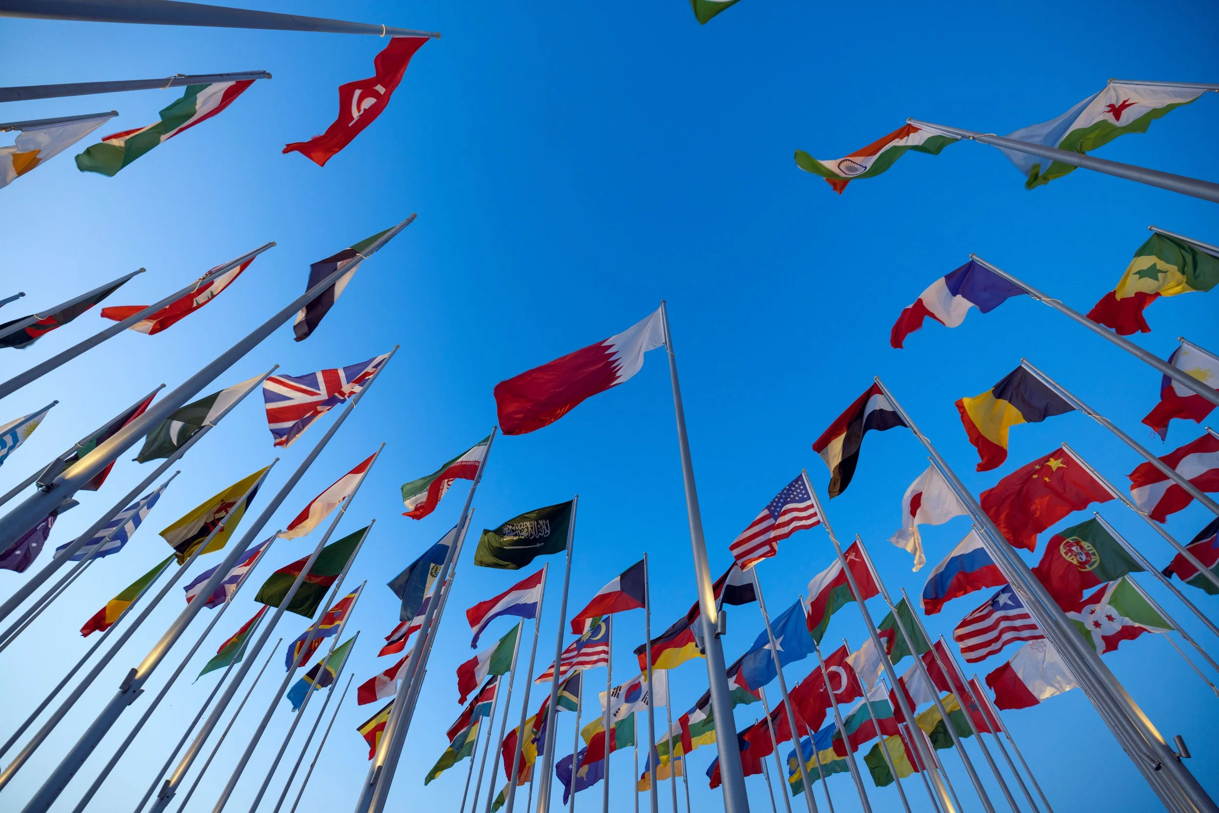 Multiple international flags flying on flagpoles against a clear blue sky.