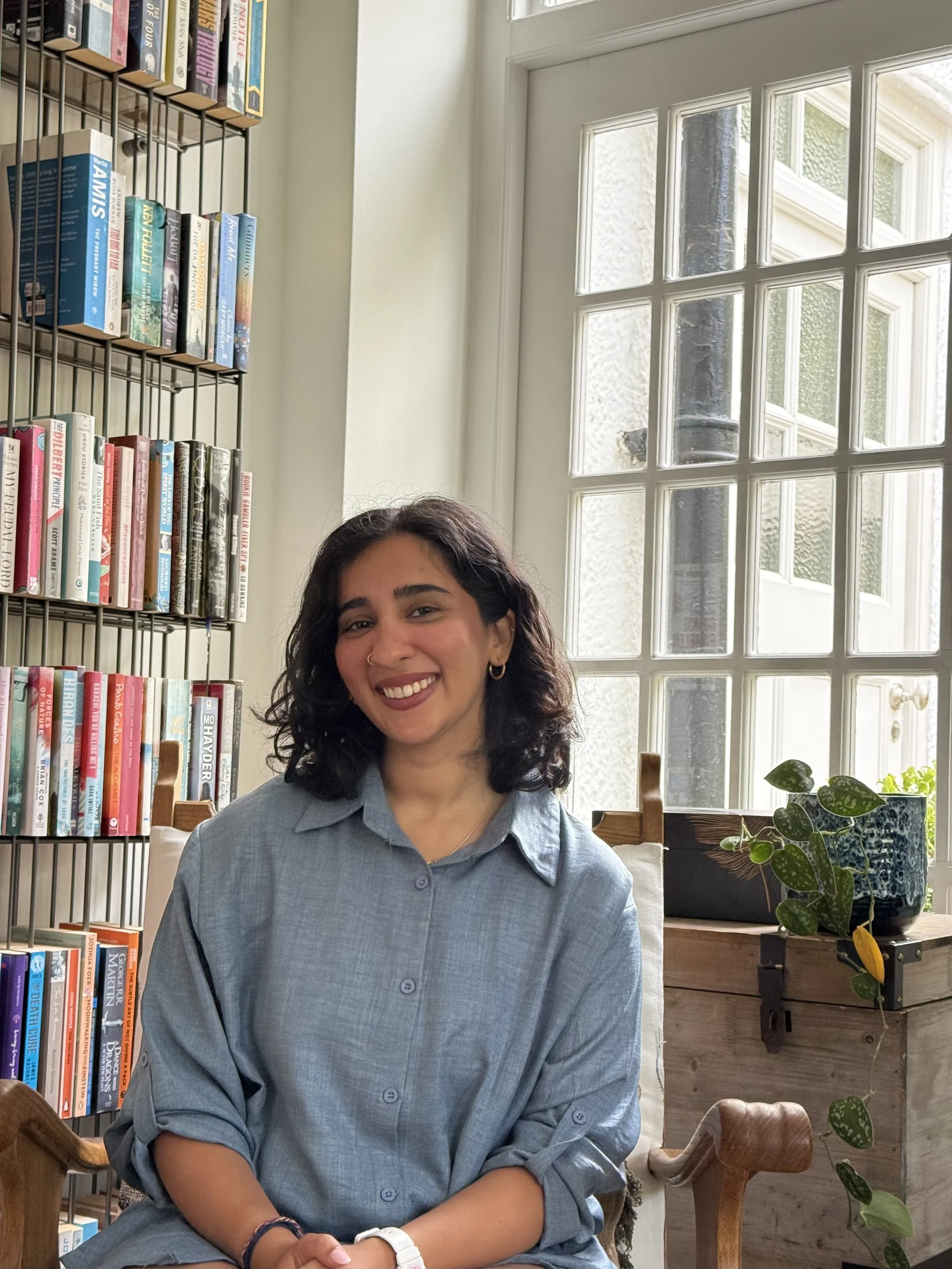 A young woman with dark wavy hair, wearing a blue button-up shirt, sitting in a cozy indoor space with a large window behind her and a bookshelf filled with books on her left. There are plants on a wooden table next to her.