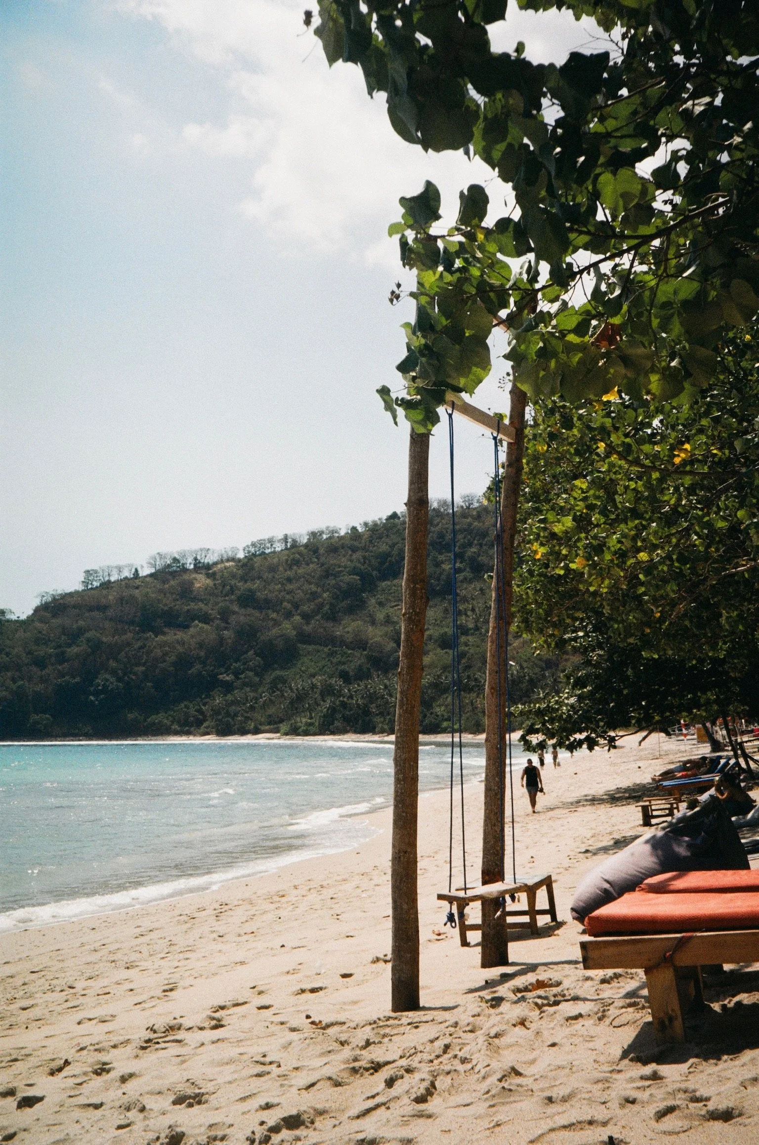 A beach scene with a swing hanging from trees on the sandy shore, boats on the sand, beach chairs, people walking along the shoreline, and a hillside in the background with trees.