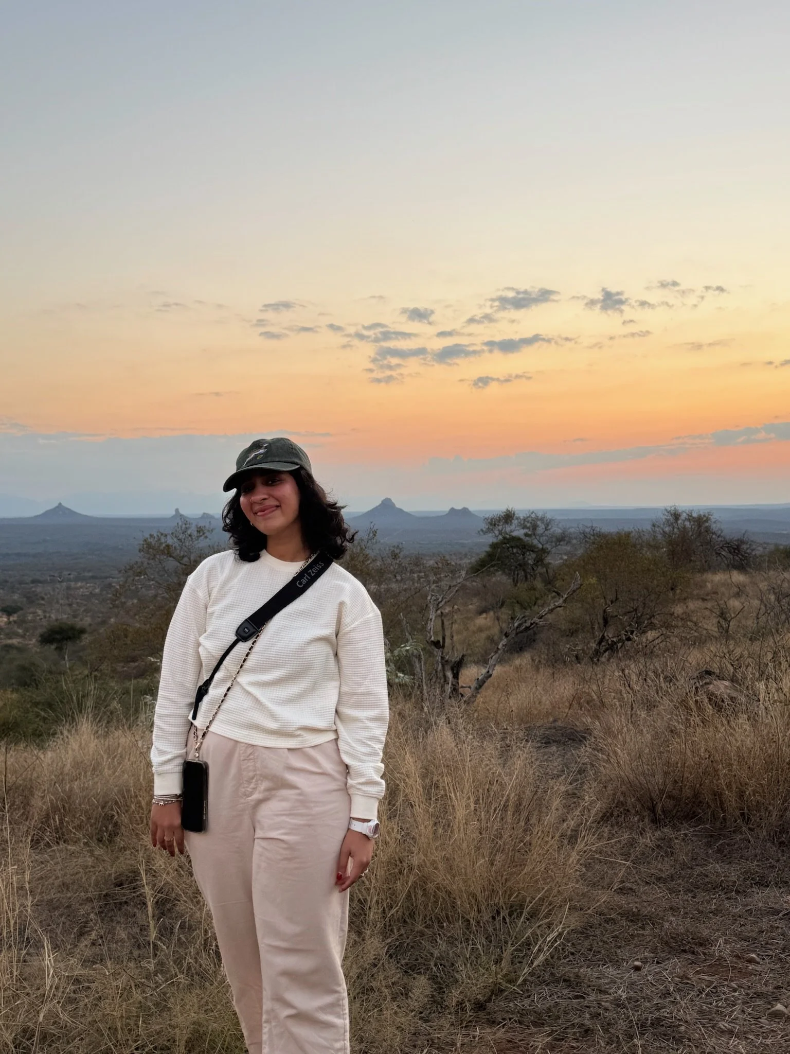 A woman in a casual outfit, wearing a beige skirt, white sweater, and a dark baseball cap, standing in a natural landscape with dry grass and sparse trees during sunset, with mountains or volcanic cones in the background.