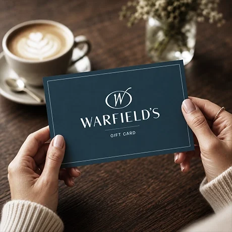 Hands holding a Warfield's gift card on a wooden table with a cup of latte and a small plant in the background.