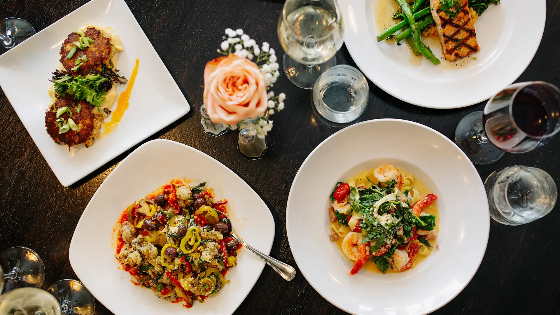 A table setting featuring various dishes, including a plate with fried food and greens, a bowl of pasta with shrimp and vegetables, a salad, and a plate of grilled meat with green beans, along with glasses of red and white wine and a small bouquet of flowers.