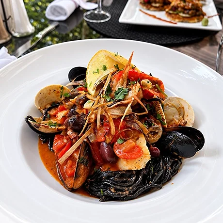 Seafood pasta with black squid ink spaghetti, shrimp, mussels, and tomatoes, garnished with lemon and herbs on a white plate.