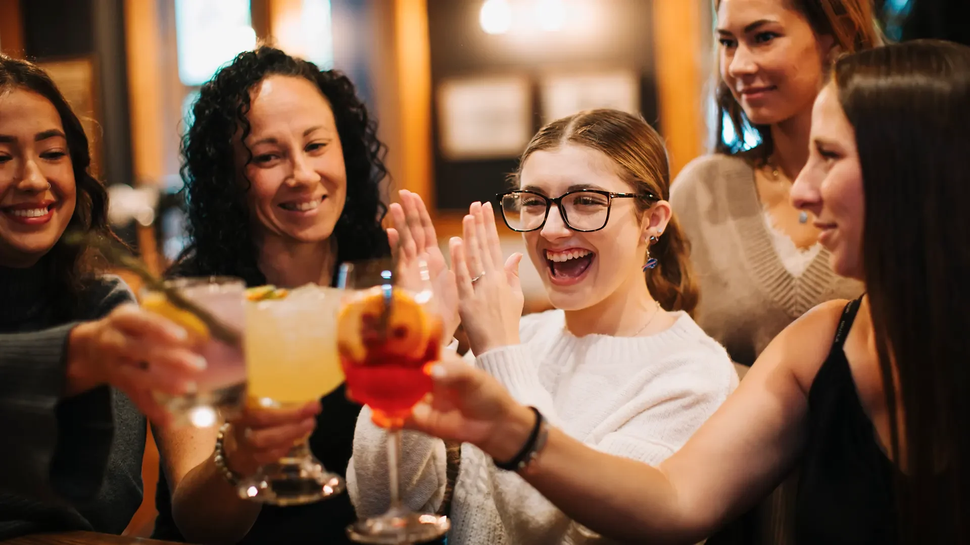 A group of women celebrating at Warfield's restaurant in Clifton Springs, NY, raising colorful cocktails in a toast, smiling and enjoying each other's company.