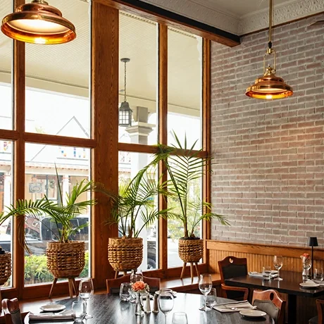 Interior of Warfield's restaurant in Clifton Springs, NY, with wooden-framed large windows, potted plants, hanging pendant lights, brick wall, and a table set with glasses and plates.