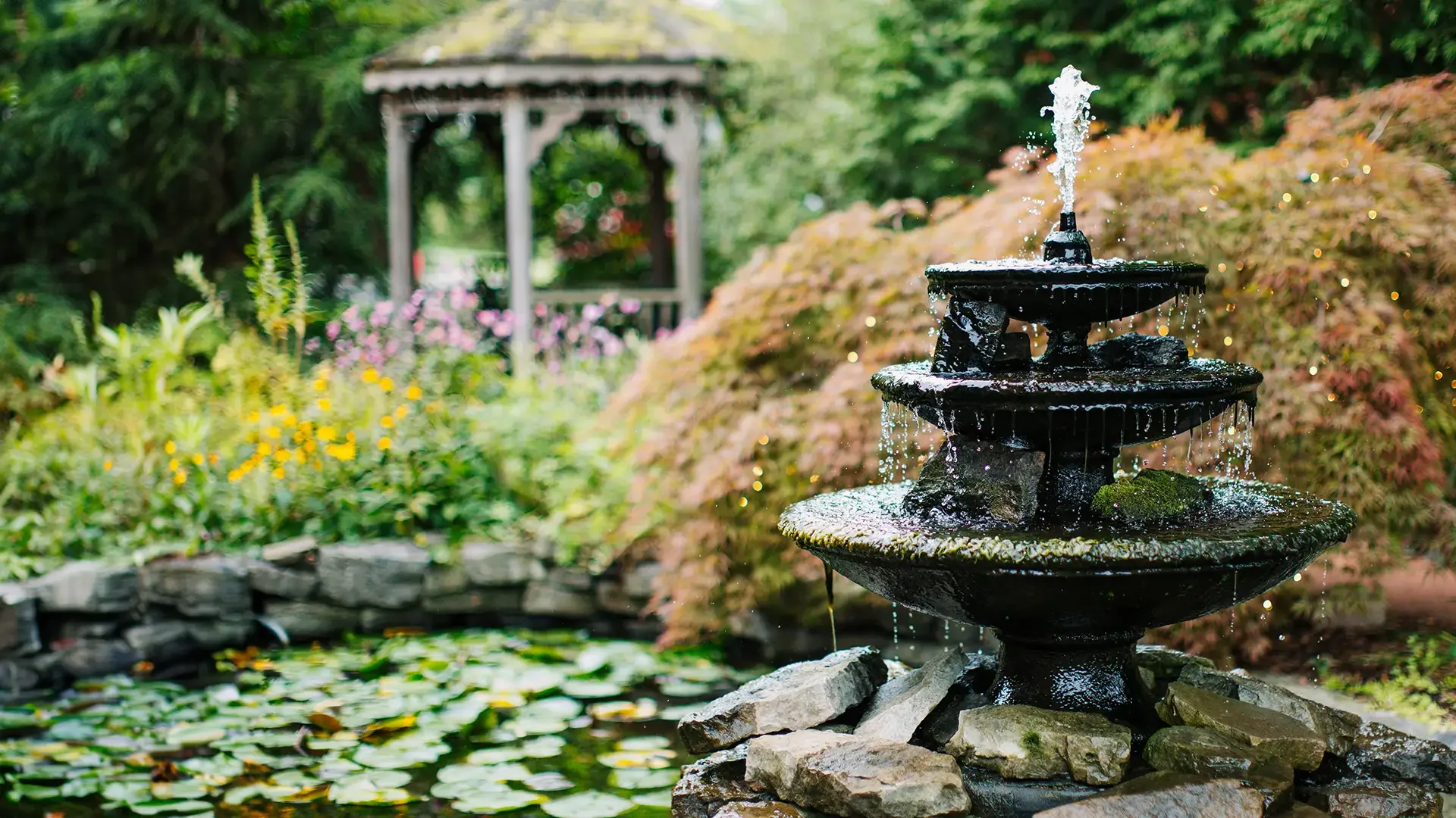 A black, tiered water fountain with water spraying from the top, surrounded by stones and lush greenery in the English garden at Warfield's restaurant in Clifton Springs, NY.
