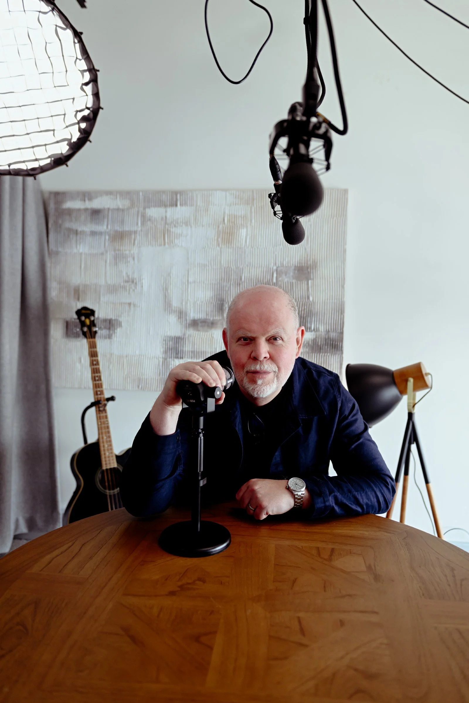 A middle-aged man with a bald head and gray beard sitting at a wooden table, holding a microphone, with studio lighting equipment and a guitar in the background.