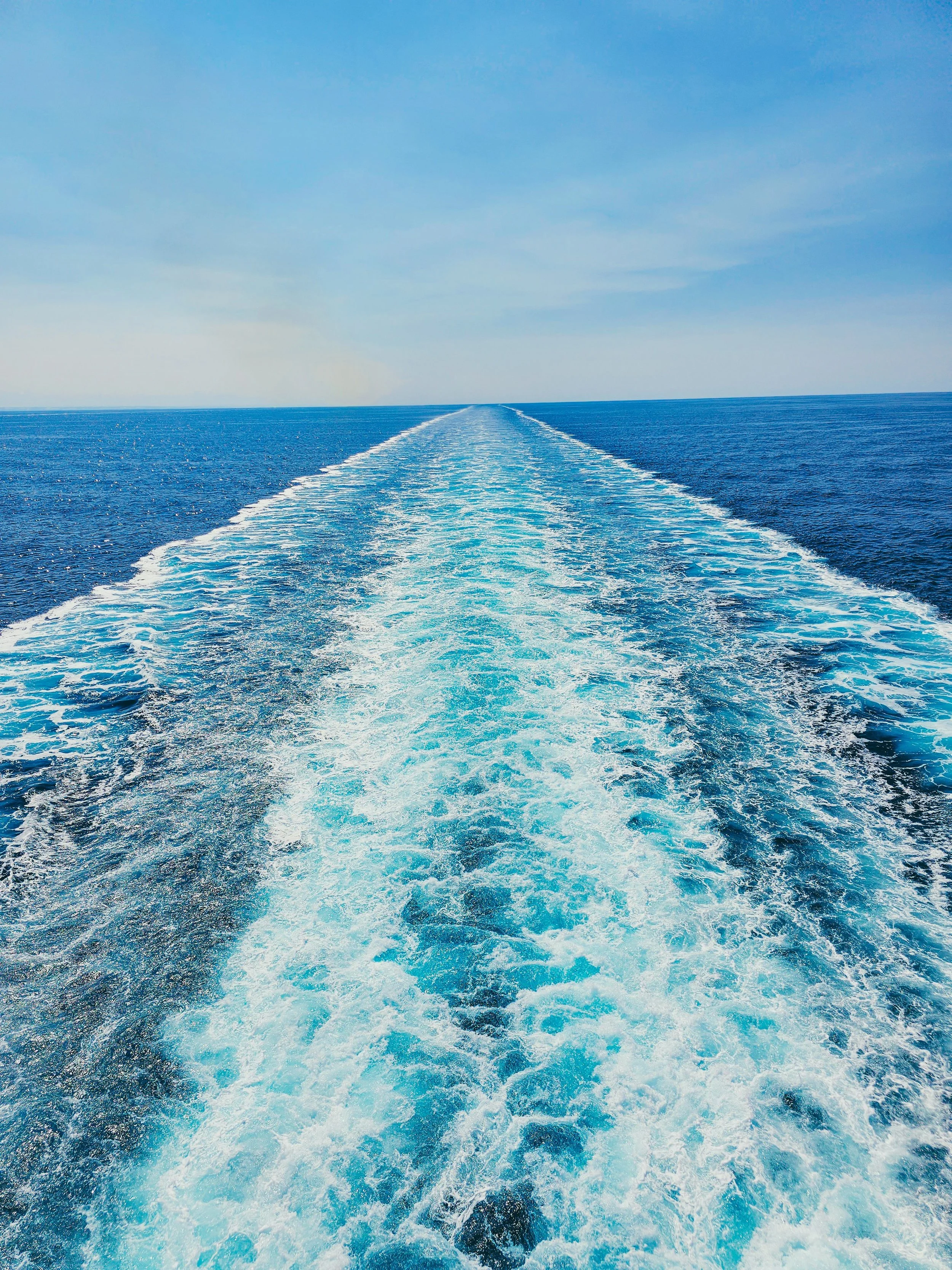 View of the ocean with a boat's wake creating a foamy trail in the water, stretching towards the horizon under a blue sky.