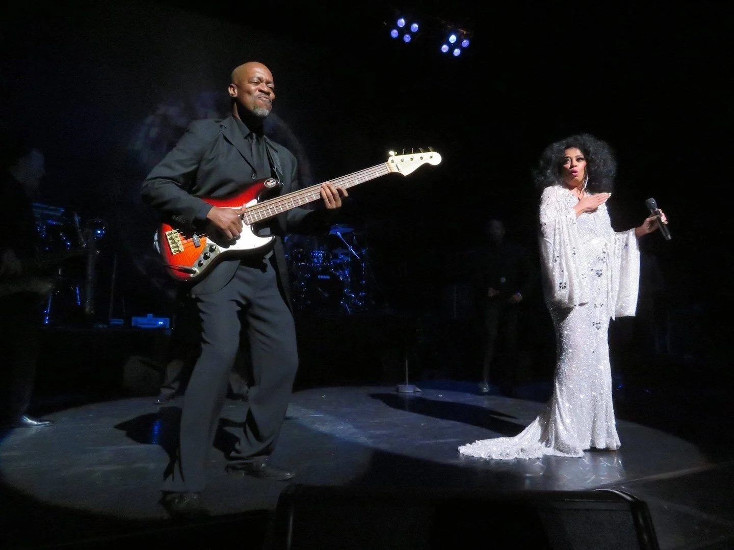 A man playing an electric guitar on stage and a woman singing into a microphone, both performing under stage lights with dark background.