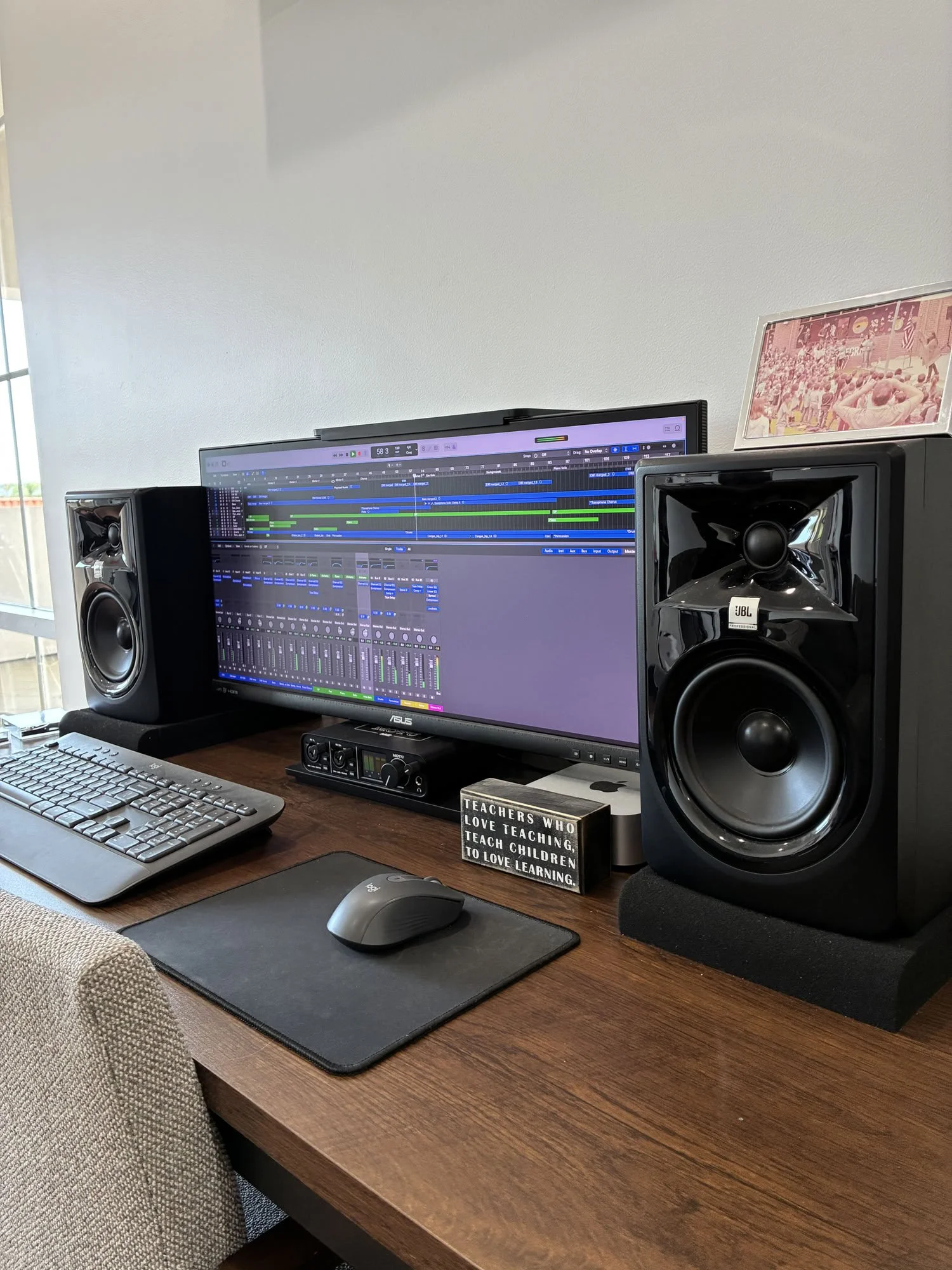 A computer workstation with a dual monitor setup, speakers, keyboard, mouse on a wooden desk, a framed photograph, and a decorative sign that says 'Teachers who love teaching, teach children to love learning'.