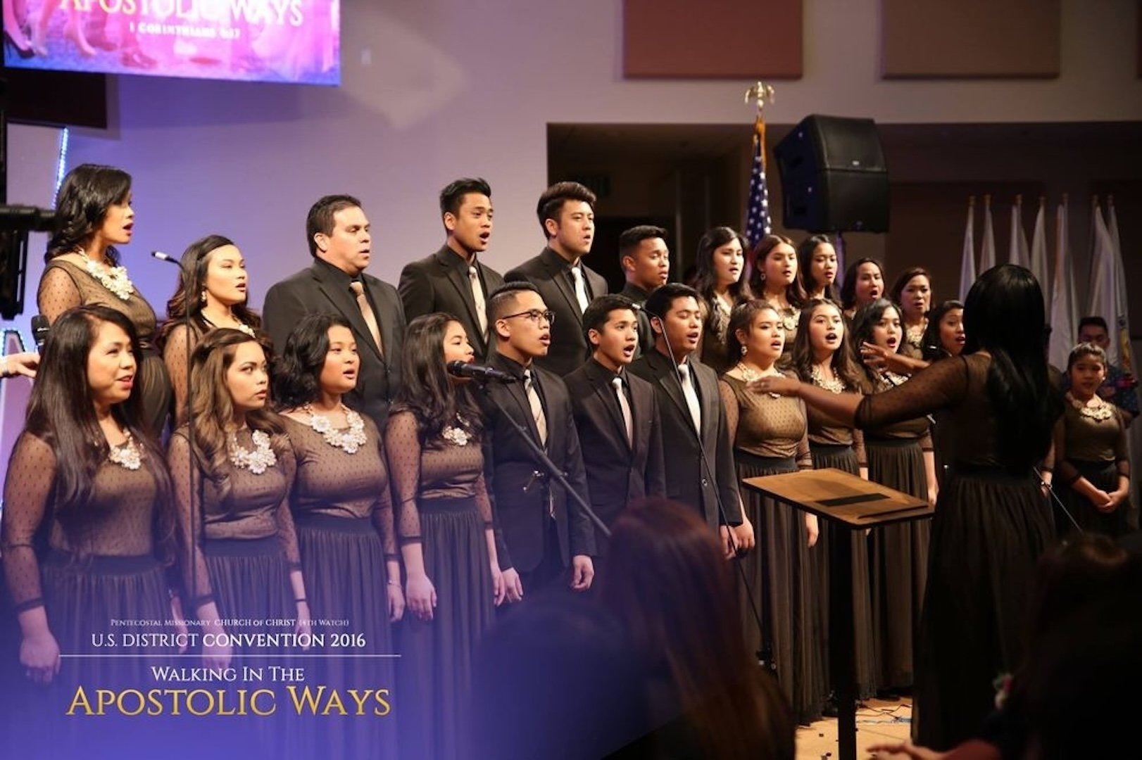 A choir group performing on stage at a religious event, directed by a conductor woman, with a banner on the bottom reading 'U.S. District Convention 2016 Walking in the Apostolic Ways'.