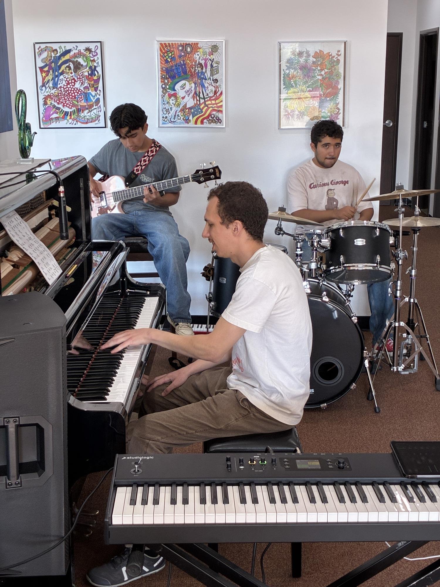 Group of three young men playing musical instruments indoors. One playing piano, one on drums, and one with guitar. Art posters on the wall behind them.