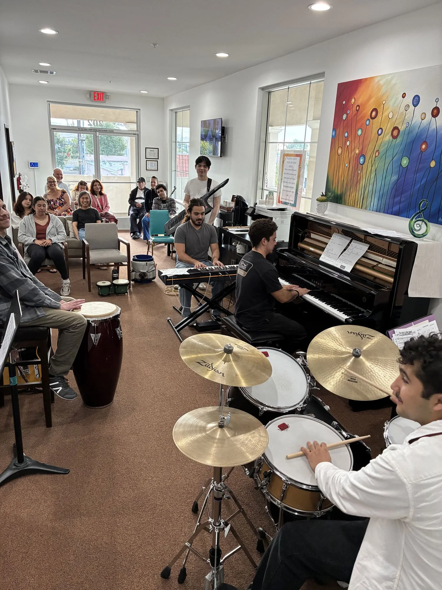 A group of people seated inside a room with white walls and large windows, watching a live musical performance. The performers include a pianist, a keyboard player, and a drummer, with a colorful abstract painting on the wall behind them.