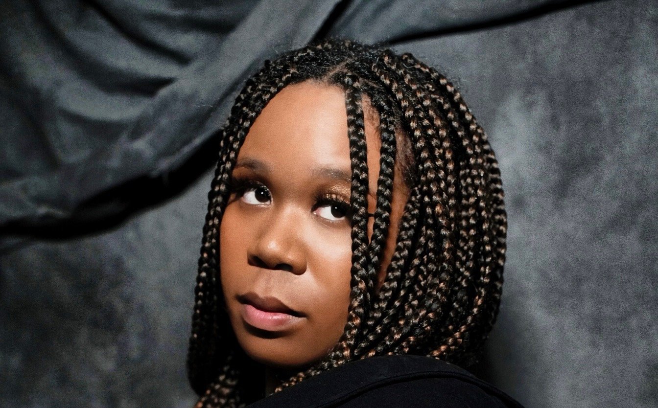 A woman with braided hair looks slightly upwards against a dark background.