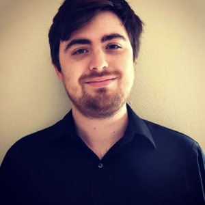 A young man with dark hair and a beard smiling, wearing a dark collared shirt, standing against a light-colored wall.