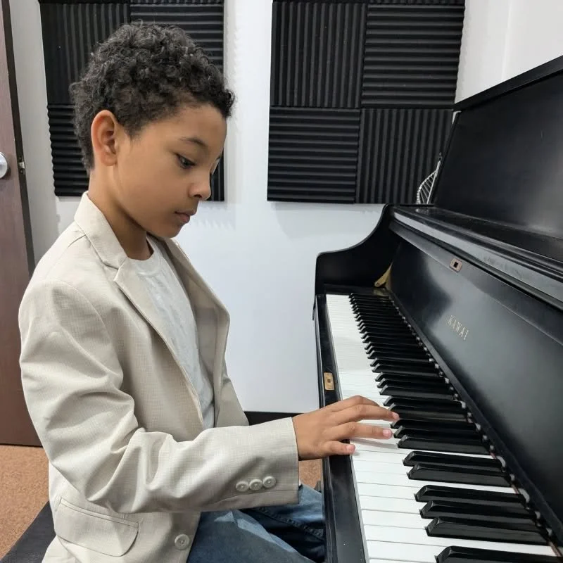 A young boy with curly hair wearing a beige blazer, white shirt, and jeans playing a black grand piano in a music studio with black acoustic foam panels on the walls.