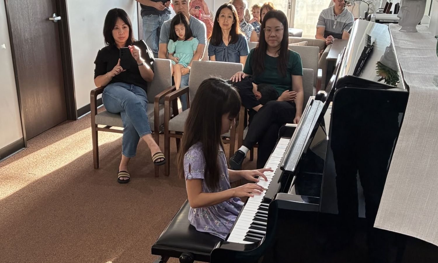 A young girl playing the piano in front of an audience of adults and children seated in chairs.