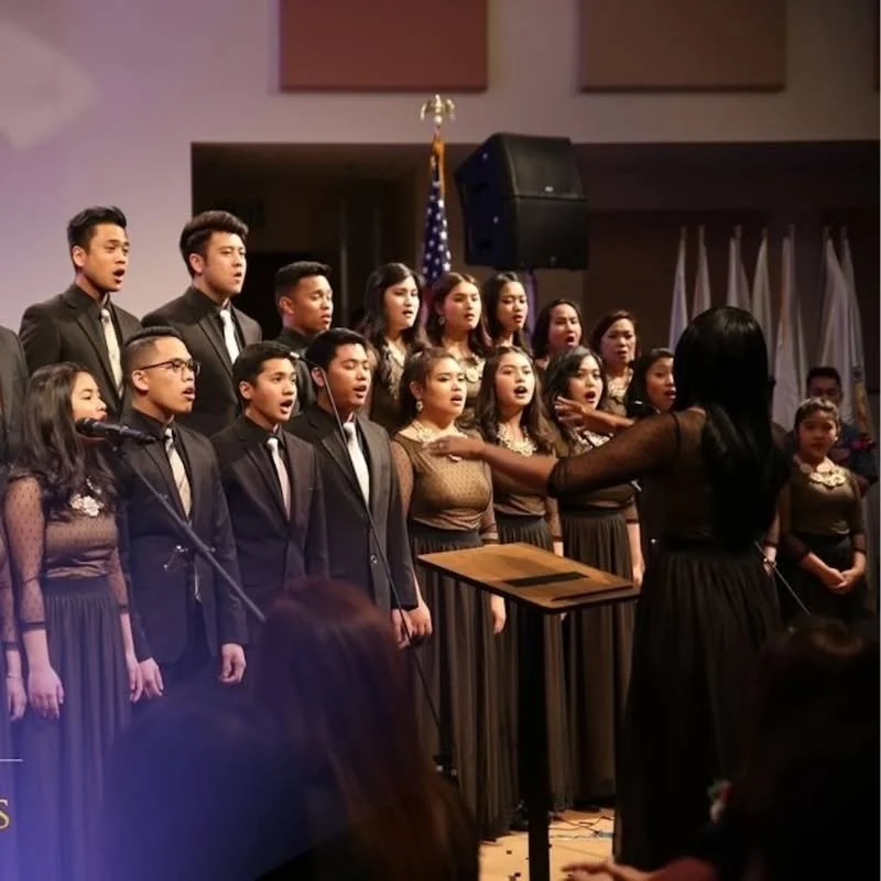 A choir group performing on stage at a religious event, directed by a conductor woman, with a banner on the bottom reading 'U.S. District Convention 2016 Walking in the Apostolic Ways'.