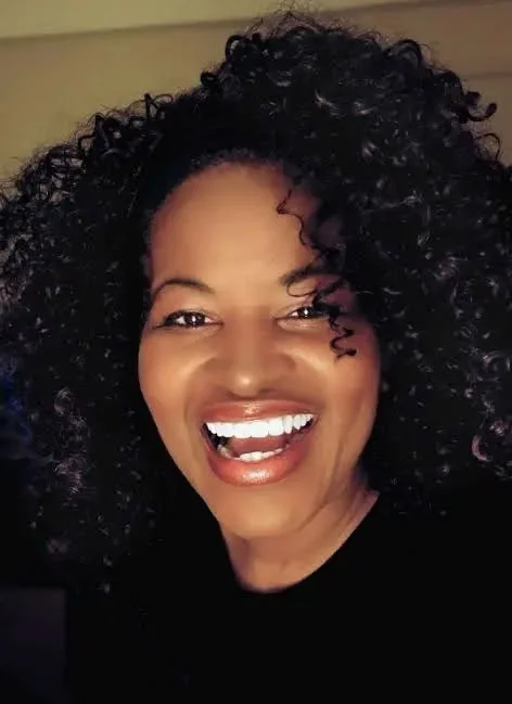 A woman with curly hair smiling and showing her teeth, against a plain background.